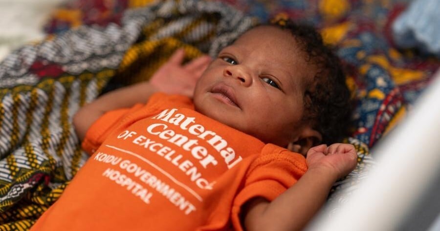 A baby lays on top of a color bedsheet. She wears a bright orange onesie with text reading 