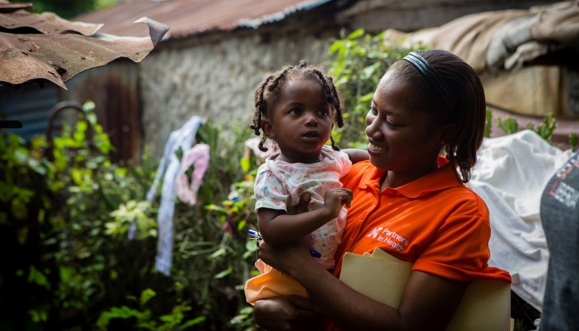 A caregiver in a bright orange PIH shirt carries a small child.