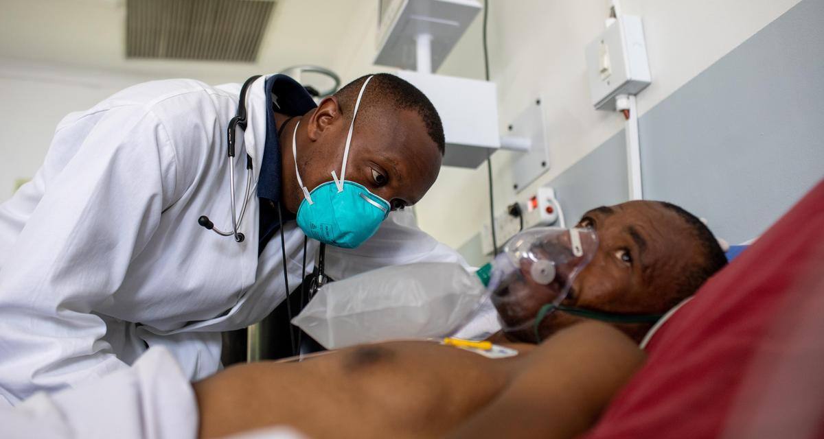A doctor wearing a lab coat and face mask leans in and checks on a man laying in a hospital bed and oxygen mask.