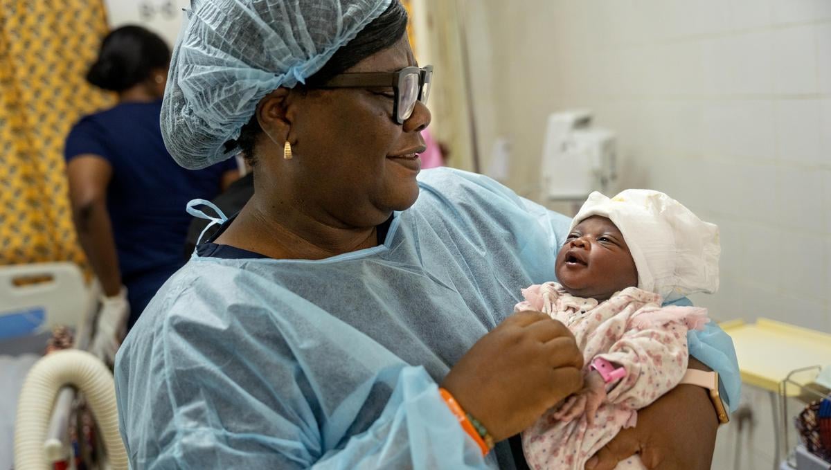A woman in personal protective equipment smiles and holds a newborn baby in her arms.